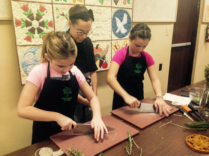 A Tree House Kitchen staff member teaches two 11-year-old girls to safely chop rosemary in a S.H.E. cooking class.