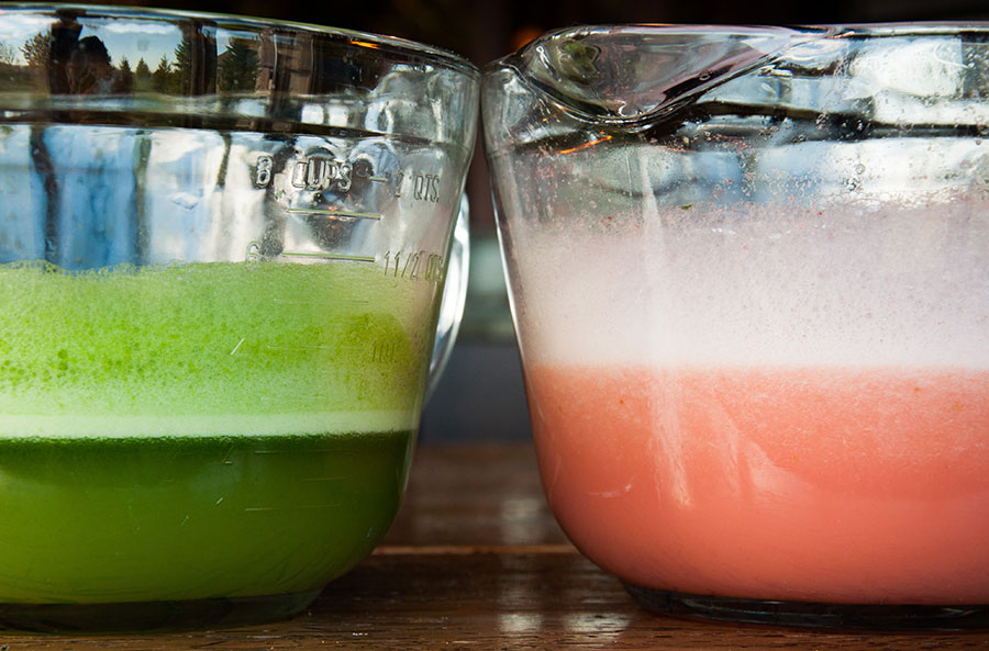 Close-up of two glass measuring cups containing edible foams—a green one created with arugula, and a pink one created with watermelon.