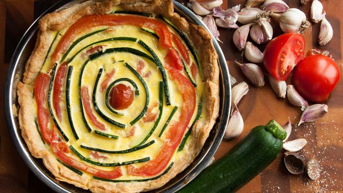 Farm vegetable and garlic crème fraîche pie, with ingredients (zucchini, tomato and garlic cloves) scattered beside it.
